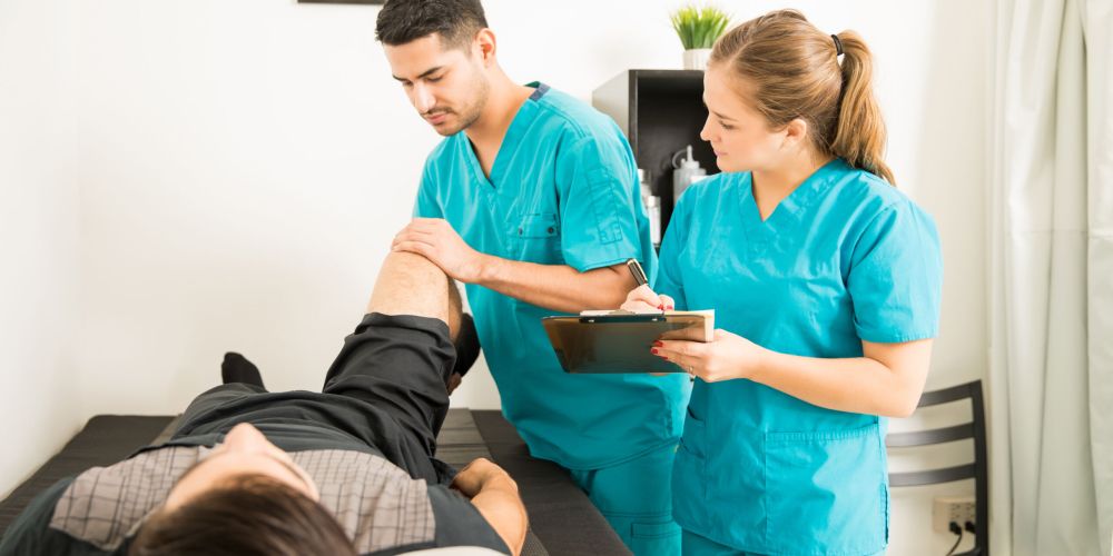 Young physiotherapist examining customer's leg while colleague noting in clipboard at hospital